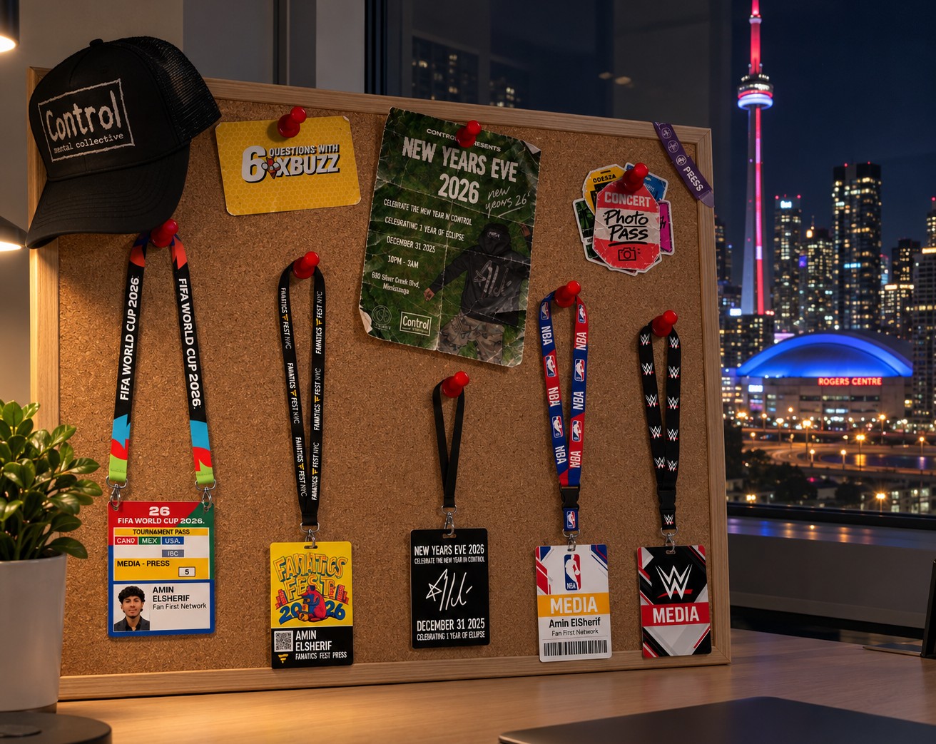 A cork board on a desk at night, pinned with press credentials, lanyards, a poster and a 6ixBuzz card, a Control cap hanging on its corner, and the Toronto skyline and Rogers Centre through the window behind it.